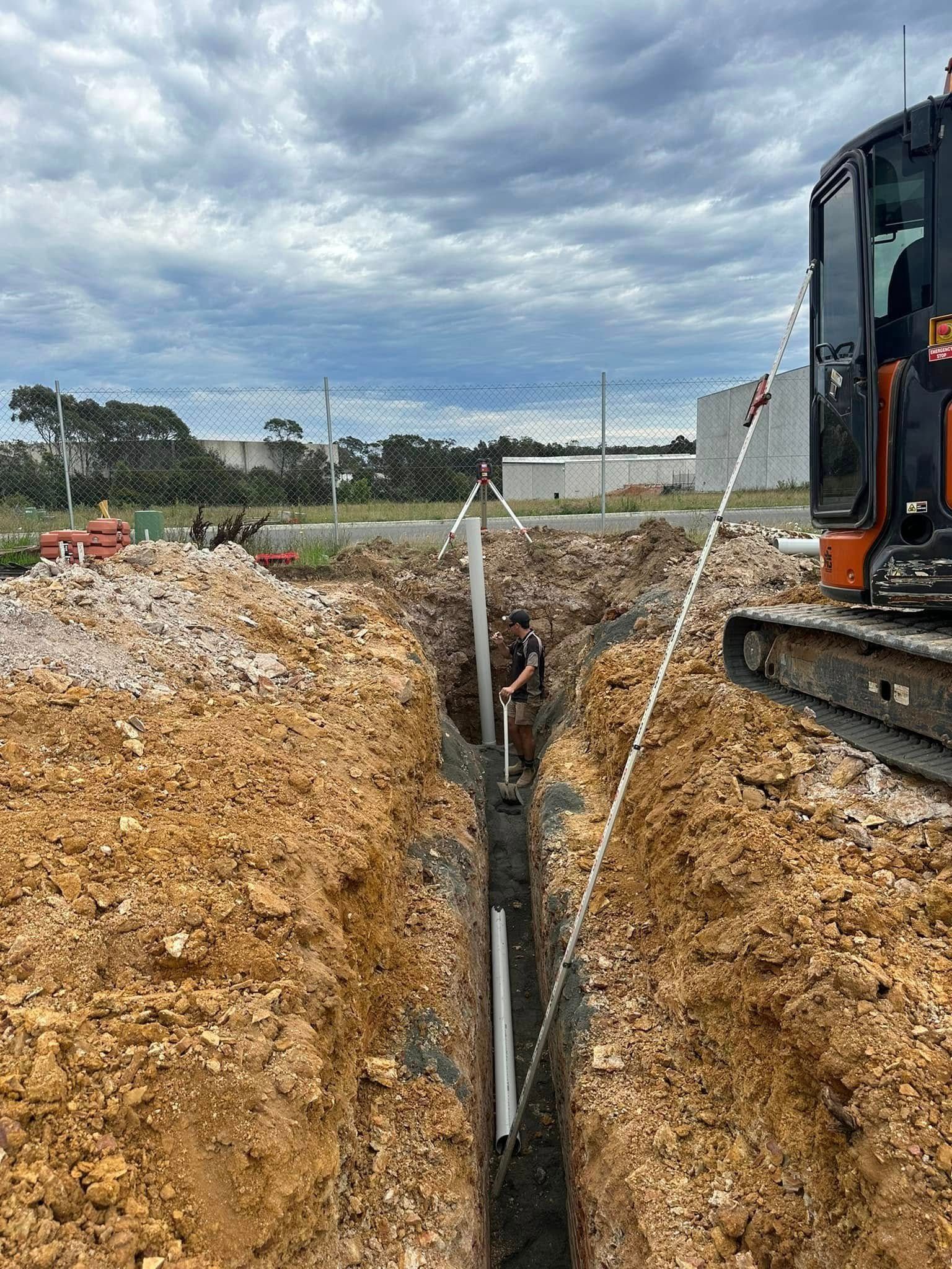 Excavator beside a deep trench lined with pipes under a cloudy sky