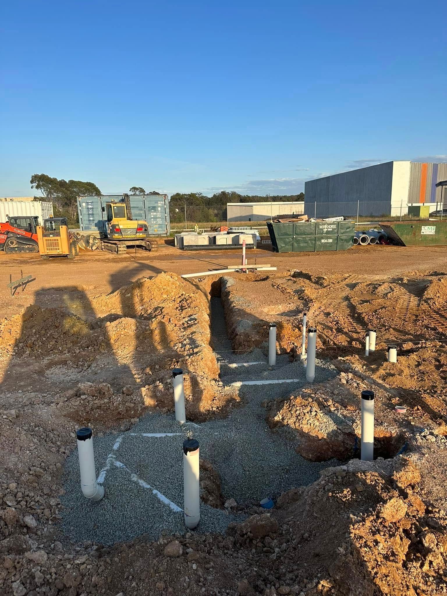 Construction site with excavated trench, exposed pipes, and heavy machinery under a clear blue sky