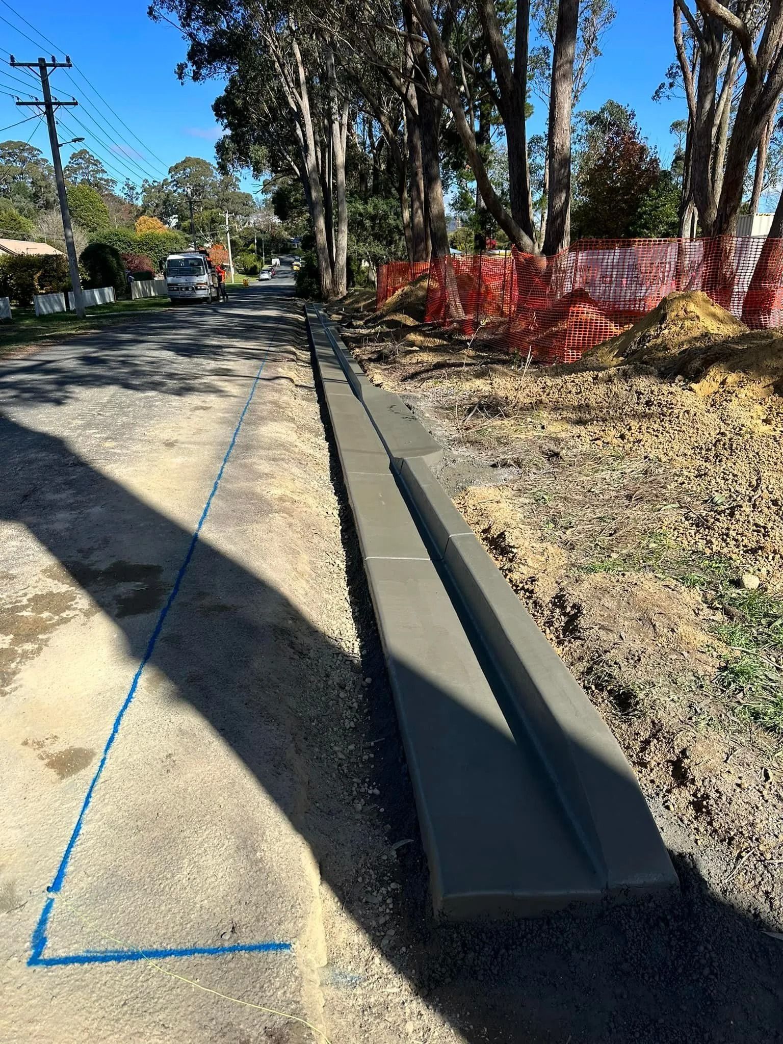 Concrete drainage channel beside a gravel path, with trees and red shrubs along a sunny street.