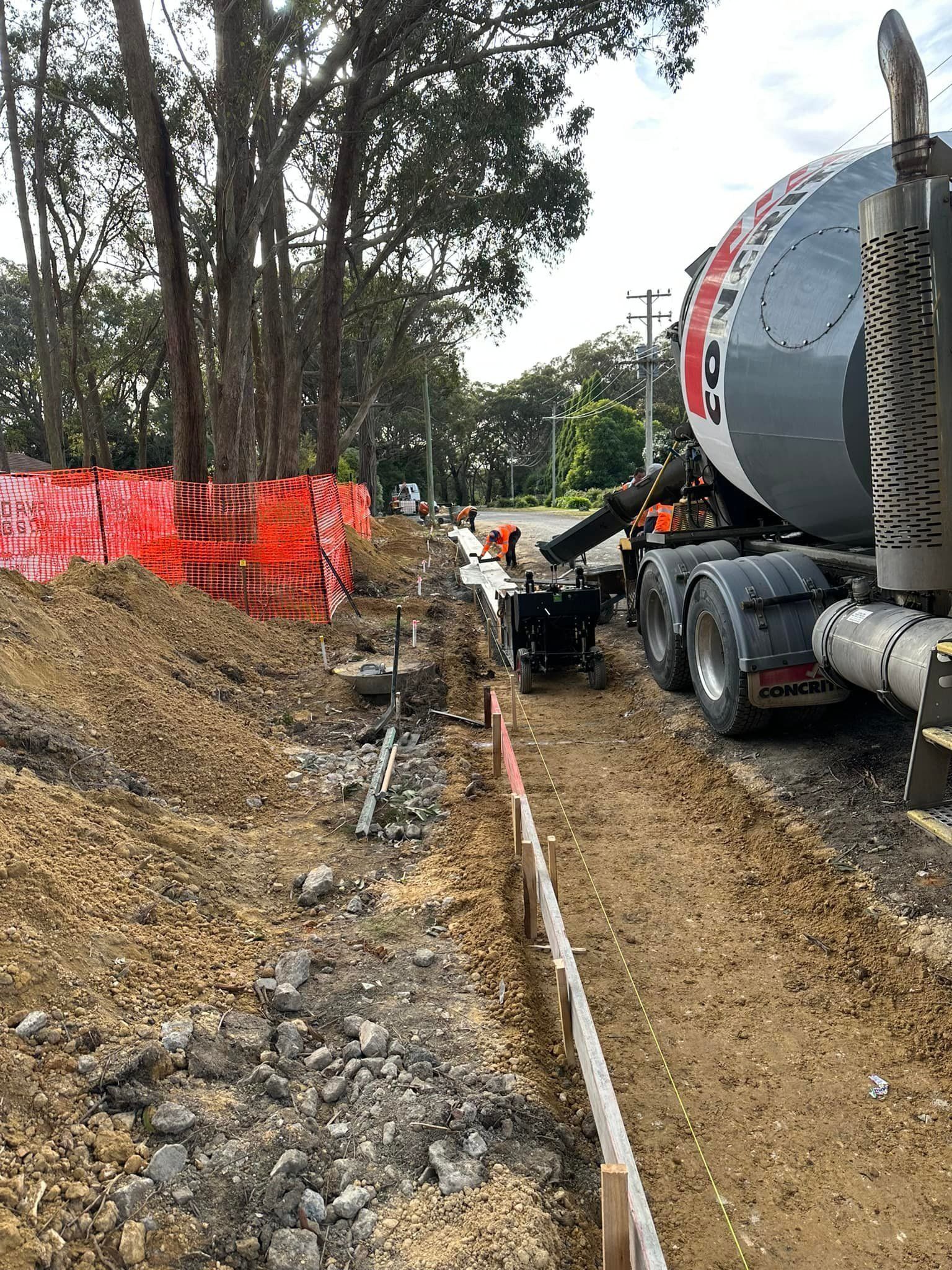 Concrete mixer truck by roadside trench, with orange safety fencing and workers near trees and gravel.