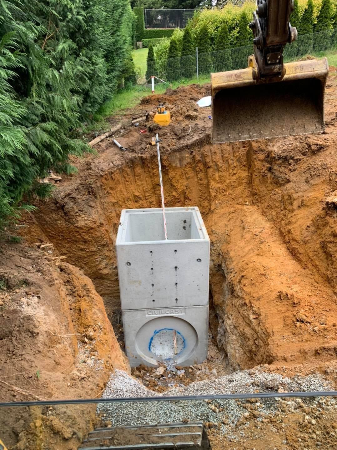 Excavator installing a concrete utility box in a deep dirt trench beside a green hedge