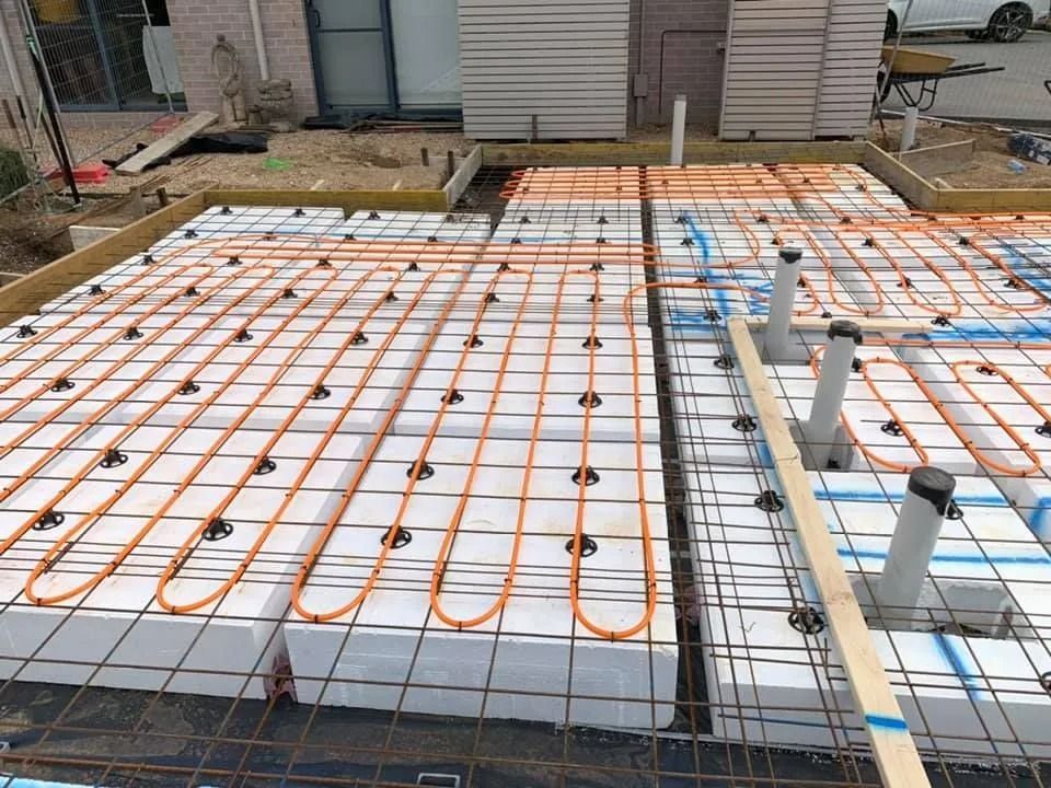 Construction site with insulated floor framing, orange tubing, and rebar grid beside a house.