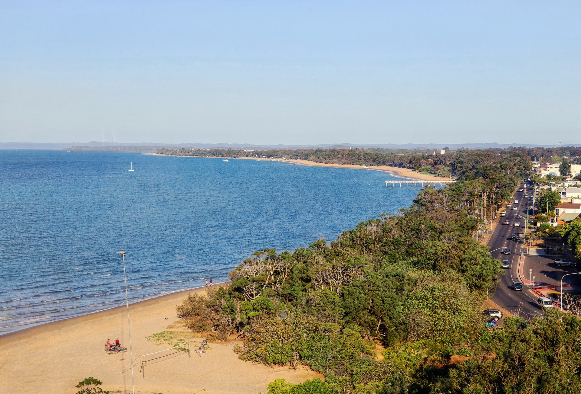 Coastal beach with blue water, sandy shore, palm trees, and a road along the coastline