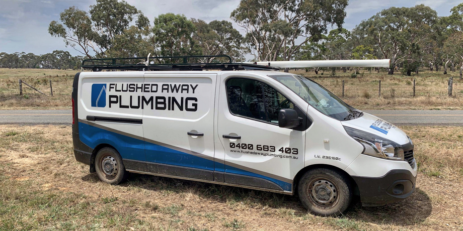 White plumbing service van parked on dirt road in a rural field, with blue “FLUSHED AWAY PLUMBING” branding.