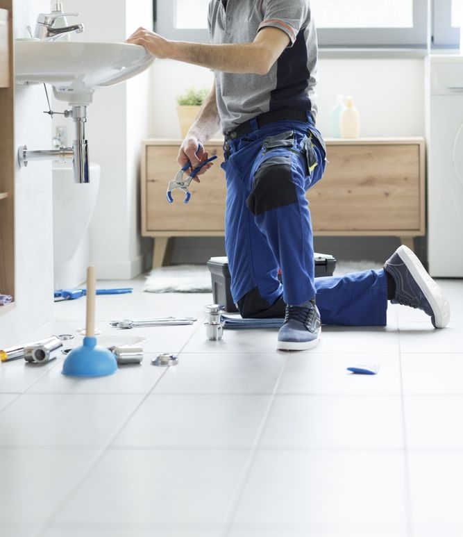 Plumber kneeling under a bathroom sink, repairing pipes on a white tiled floor with tools scattered nearby