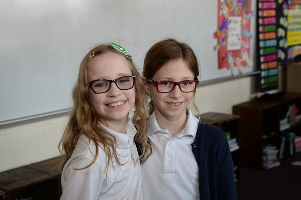Two young girls wearing glasses are posing for a picture in a classroom.