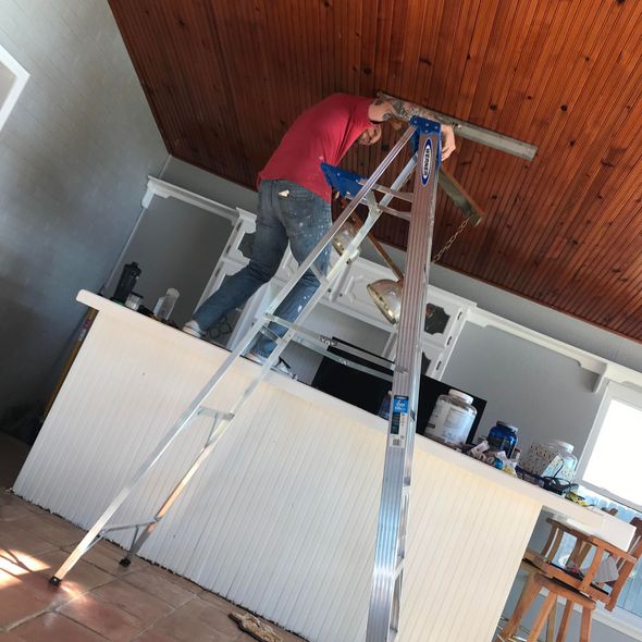 Person on a ladder working on ceiling fixture above a white counter. Interior setting, brown ceiling.