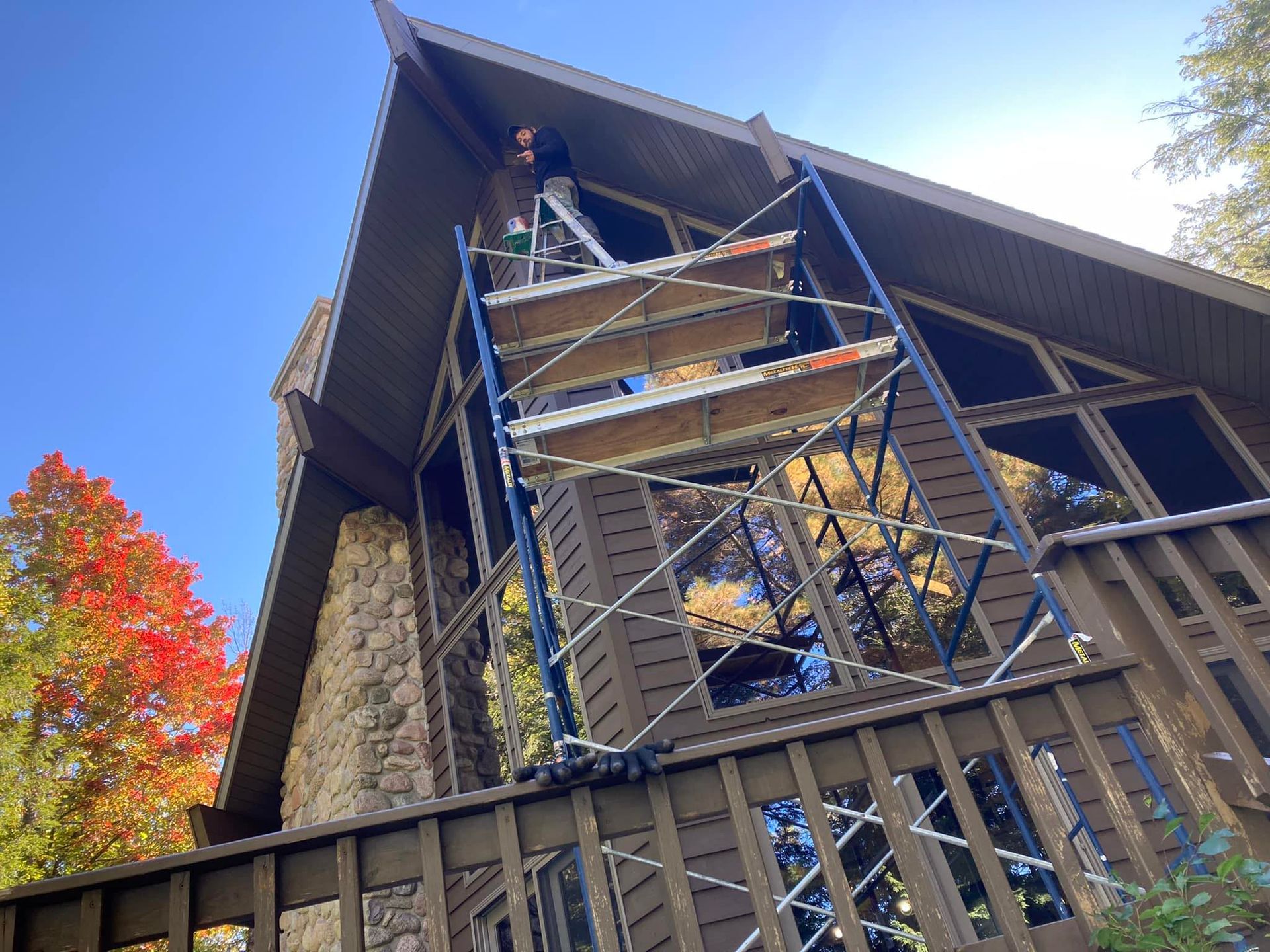 Man on scaffolding working on the exterior of a brown house with large windows; fall foliage in the background.