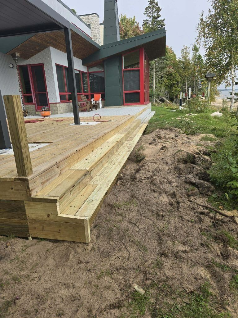 Wooden deck stairs leading down from a modern house with a green roof, next to a sandy hill.