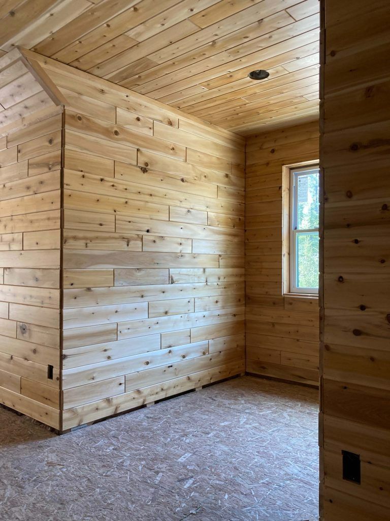 Empty room with wood-paneled walls and ceiling, a window, and exposed electrical outlets.