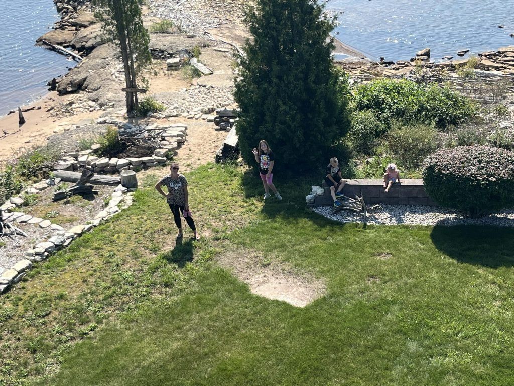 People by water. One person standing, others sitting near a brick wall. Green grass and trees visible.