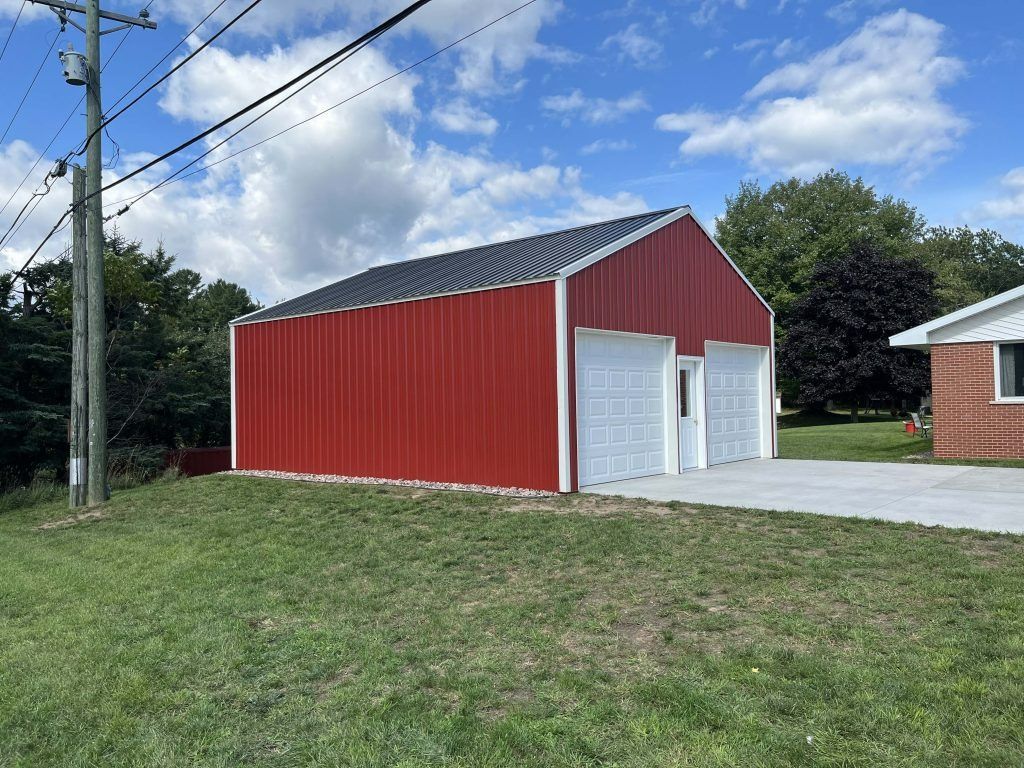 Red metal garage with white doors, black roof, and concrete driveway on a grassy hill, under a partly cloudy sky.
