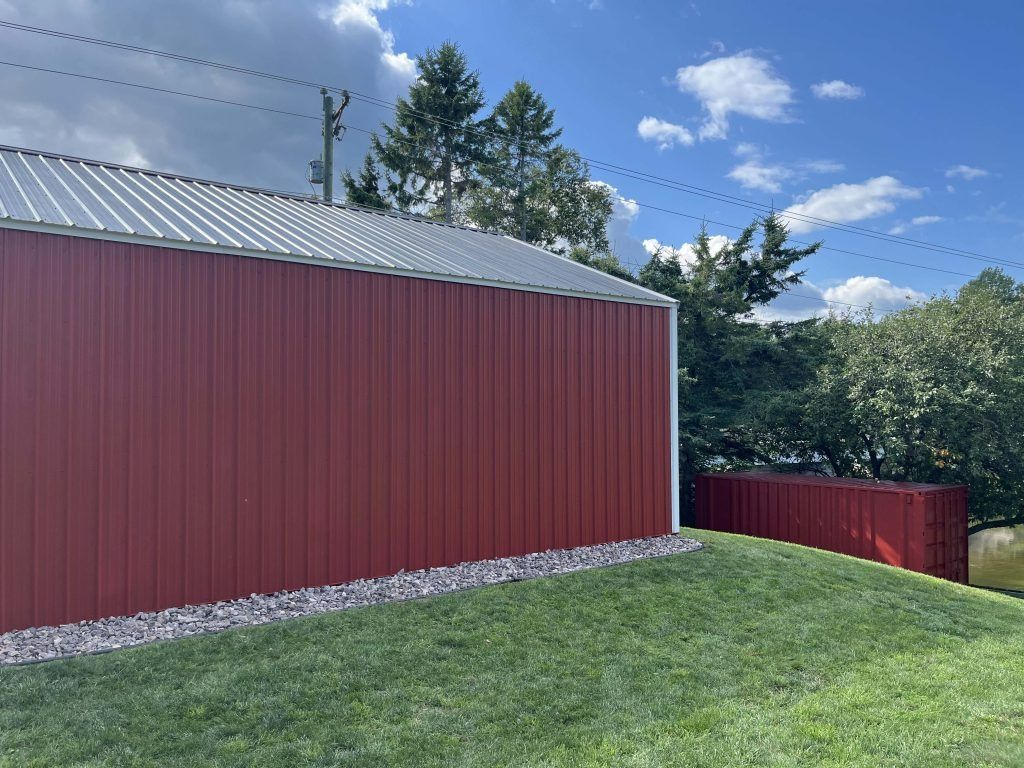 Red metal-sided building with silver roof, green lawn, gravel border, blue sky with clouds.