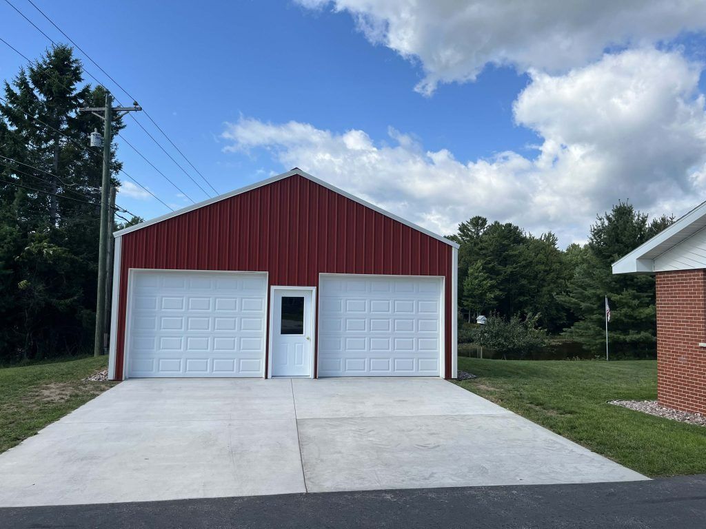 Red metal garage with two white garage doors, small door, concrete driveway, and blue sky.