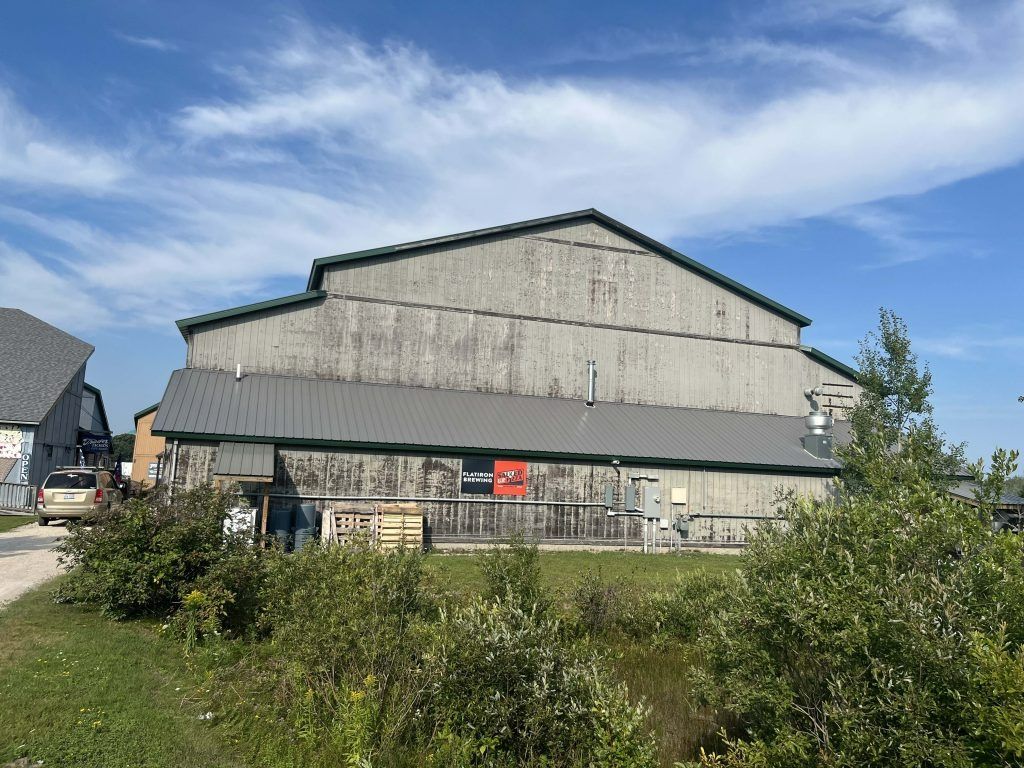 Large, weathered gray barn with a green roof against a blue sky, viewed from a grassy area.