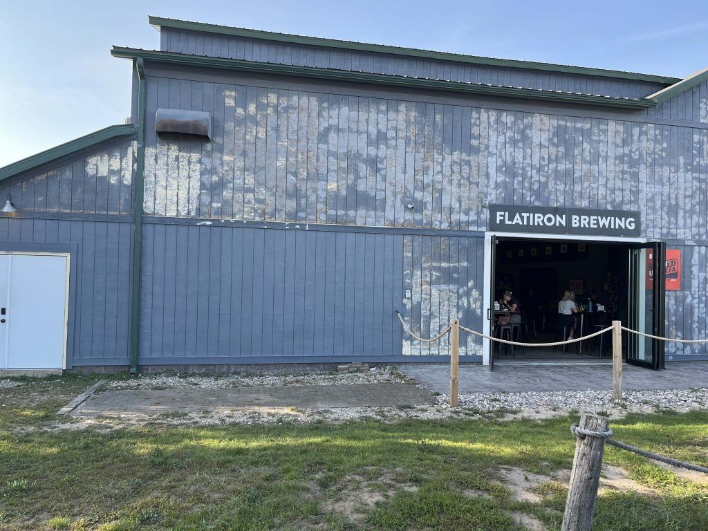FlatIron Brewing building with open doors, weathered blue paint, and a grassy area.