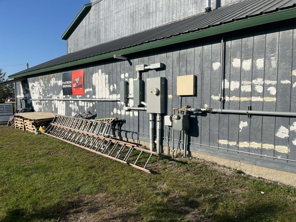 Exterior of a weathered building with a ladder, electrical boxes, and wooden pallets on the grass.
