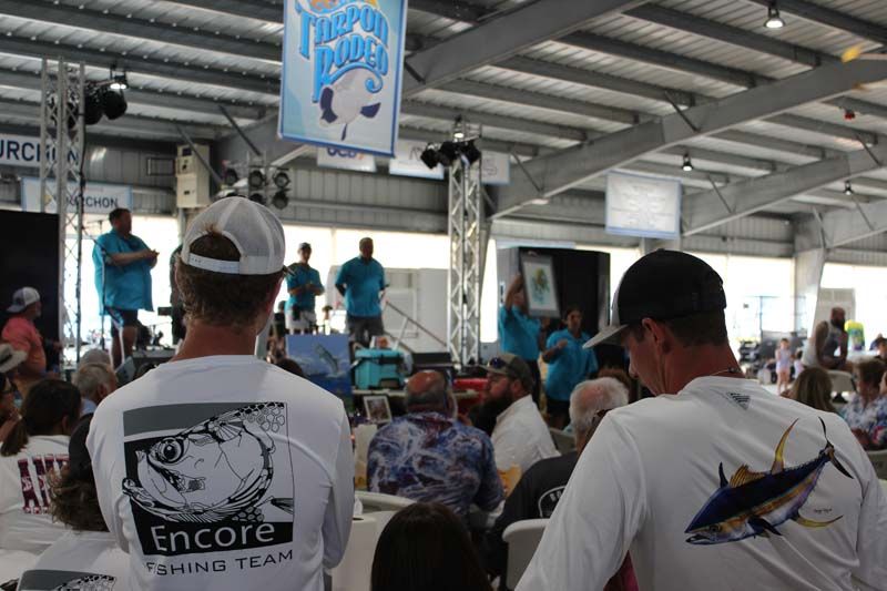 Two men wearing encore fishing team shirts are sitting in front of a crowd