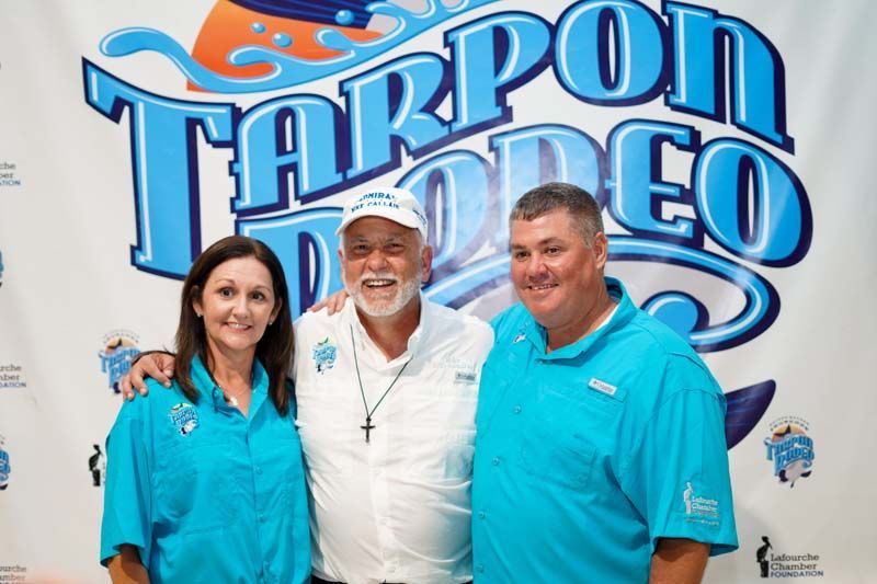 Three people are posing for a picture in front of a tarpon rodeo sign.
