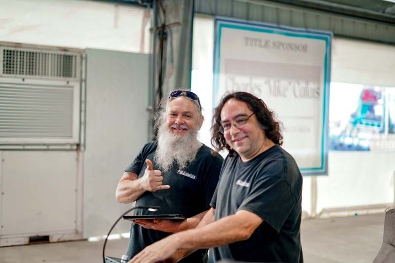 Two men with beard and long hair are standing next to each other in front of a sign.