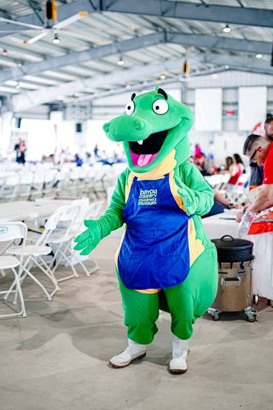 A green alligator mascot is standing in a room with tables and chairs.