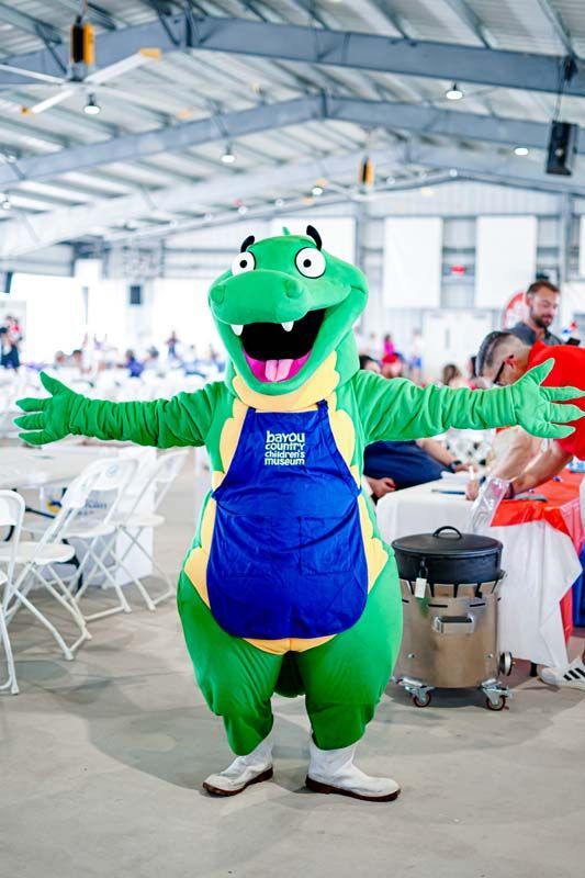 A green dinosaur mascot is standing in a room with people sitting at tables.