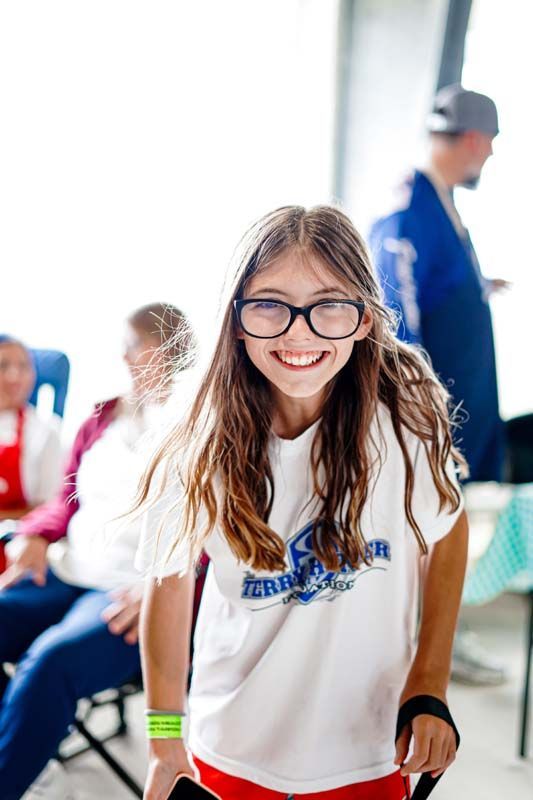 A young girl wearing glasses and a white shirt is smiling for the camera.