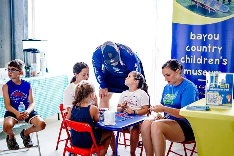 A group of children are sitting at a table in front of a sign that says bayou country children 's museum.