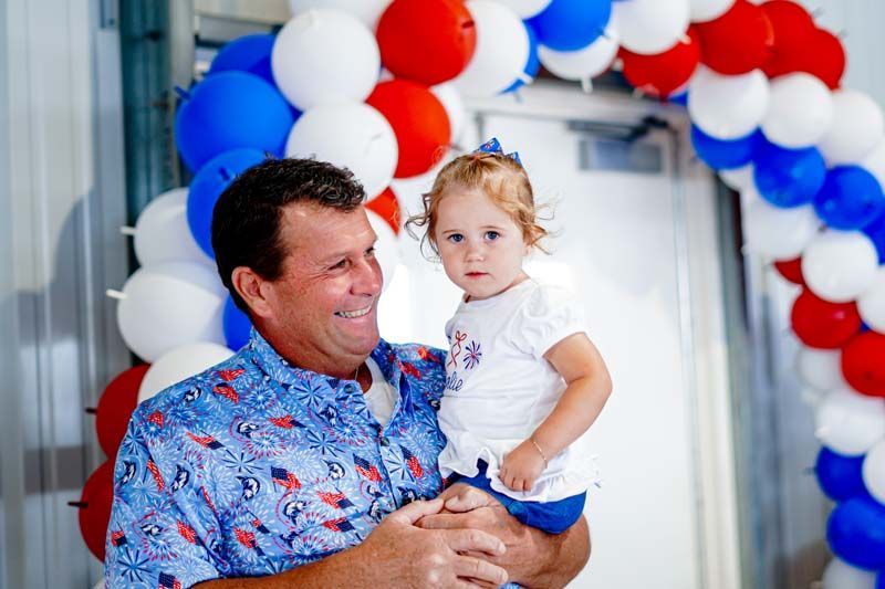 A man is holding a little girl in his arms in front of a balloon arch.