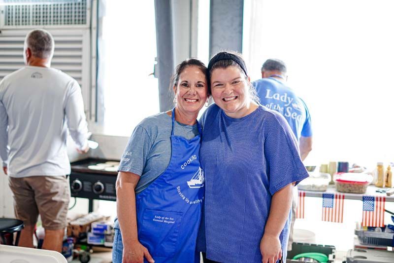 Two women wearing blue aprons are posing for a picture in a kitchen.