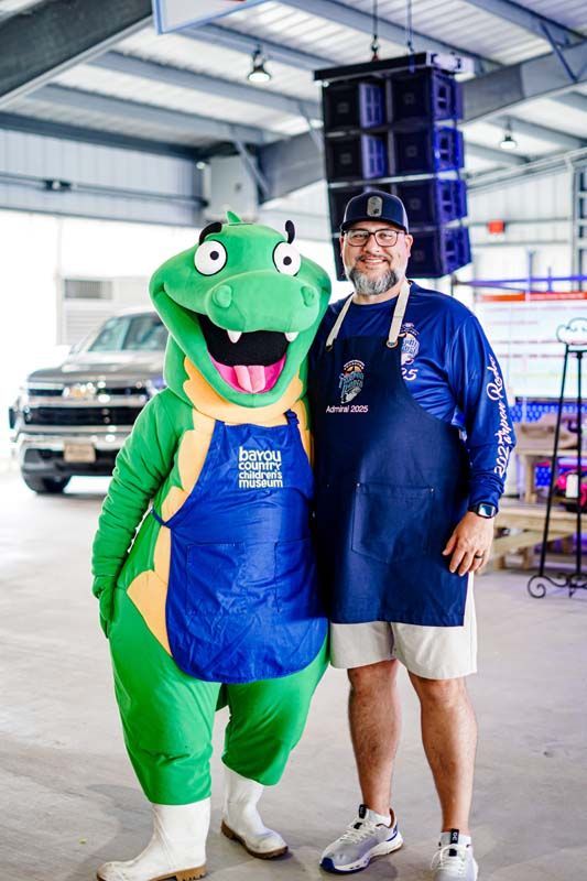 A man in an apron is standing next to a green mascot.
