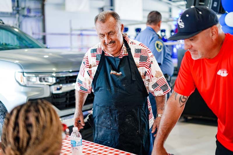 A man in an apron is standing next to a man in a red shirt.
