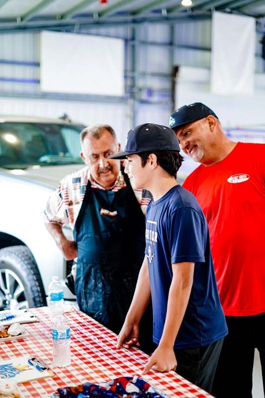 Three men are standing around a table in front of a car.