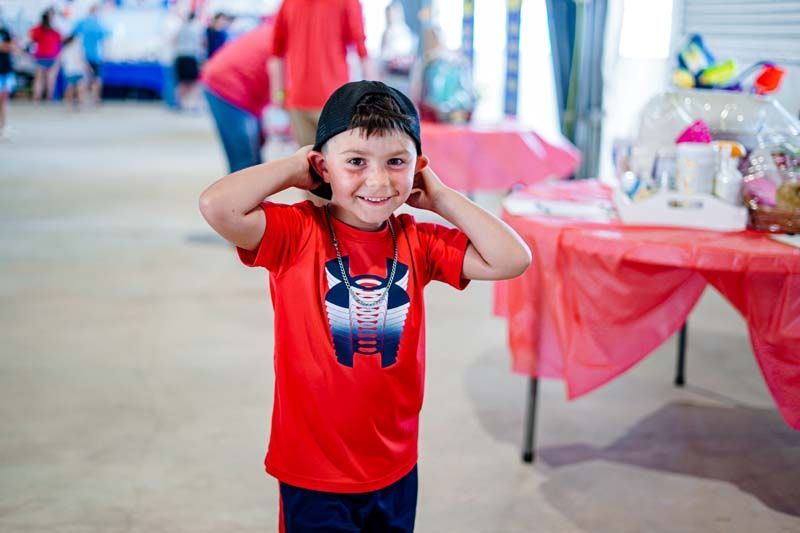 A young boy wearing a red shirt and a black hat is standing in front of a table.