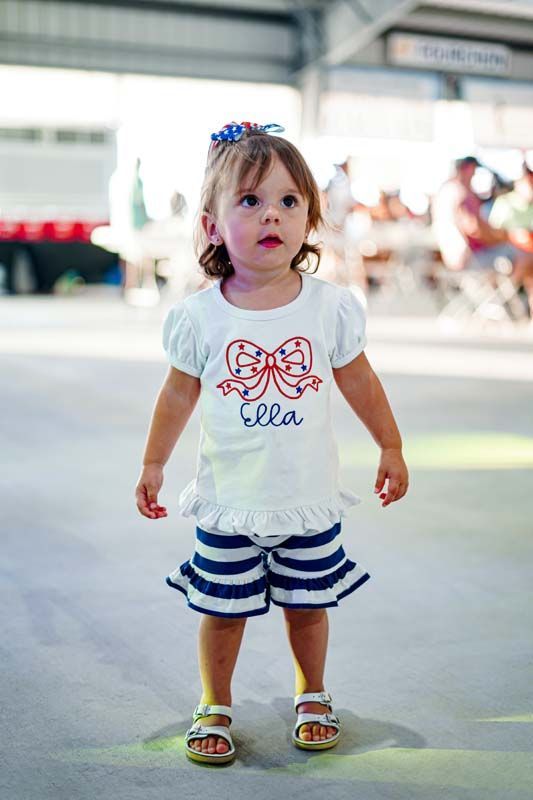 A little girl wearing a white shirt with a bow on it and striped shorts.