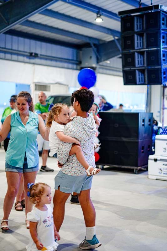 A man is holding a little girl in his arms while a group of people are dancing in a room.