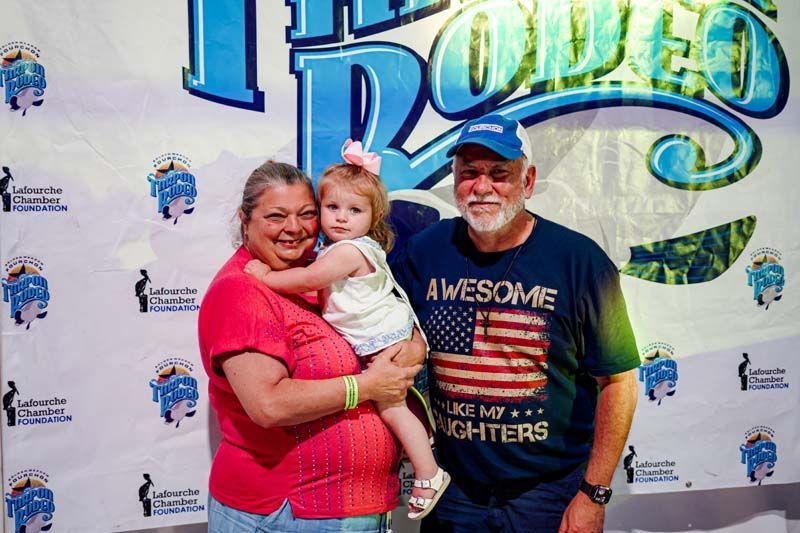 A man and woman are holding a little girl in front of a rodeo sign.