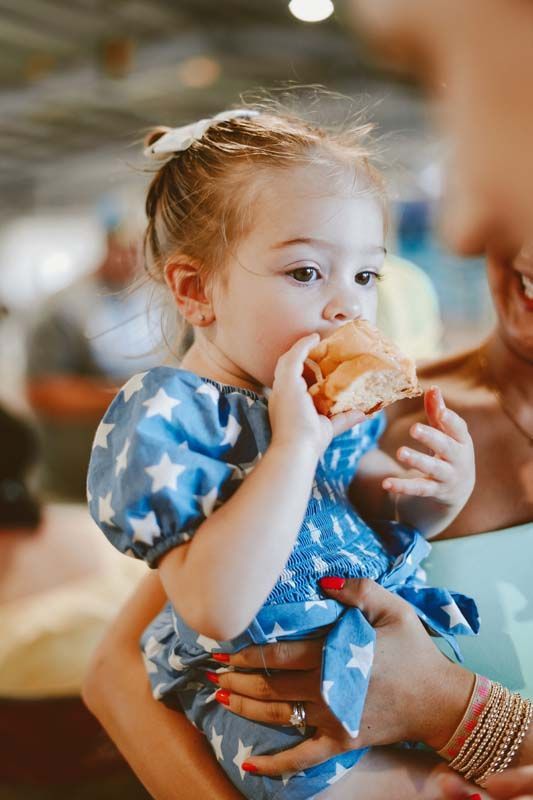 A little girl is eating a hamburger while being held by a woman.