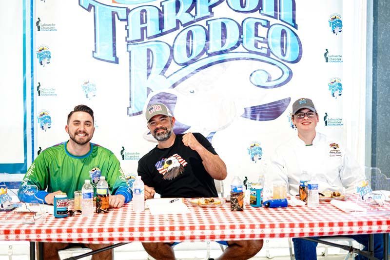 Three men are sitting at a table in front of a tarpon rodeo sign.