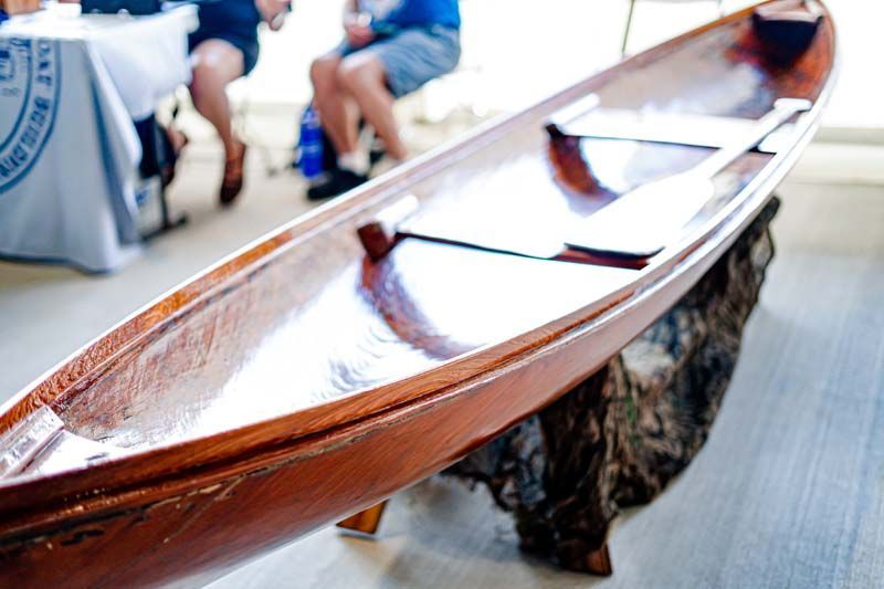 A wooden canoe is sitting on a table with people sitting at tables in the background.