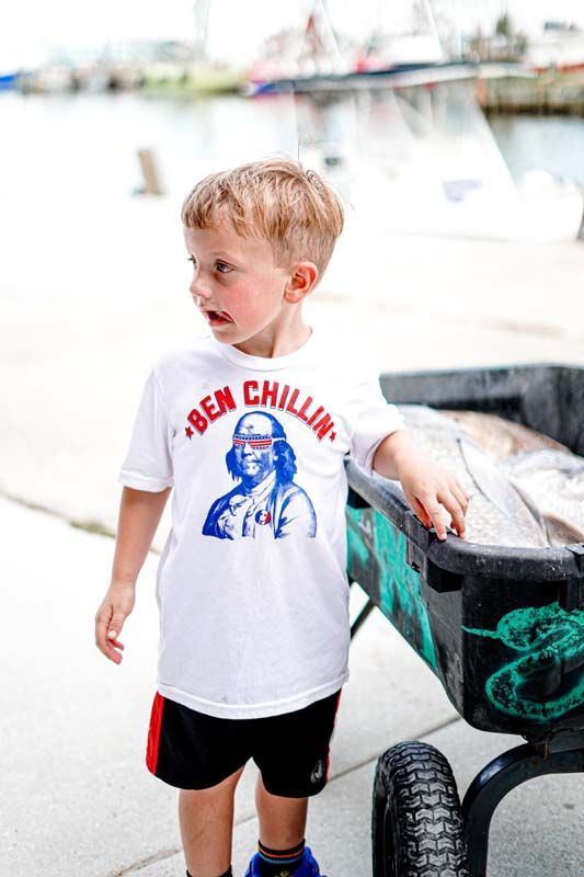 A young boy wearing a ben chillin t-shirt is standing next to a wheelbarrow.