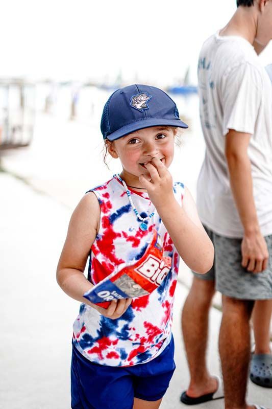 A young boy wearing a hat and a tie dye shirt is eating a bag of big mac chips.