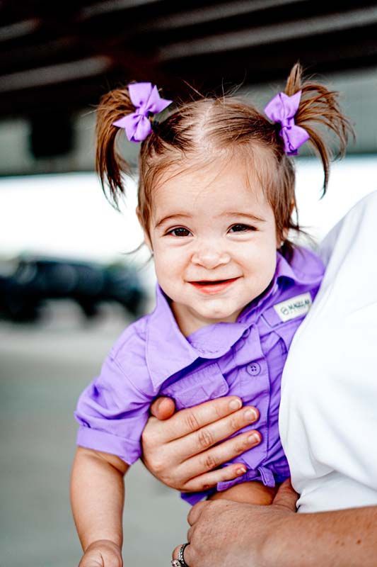 A little girl wearing a purple shirt and purple bows is being held by a woman.
