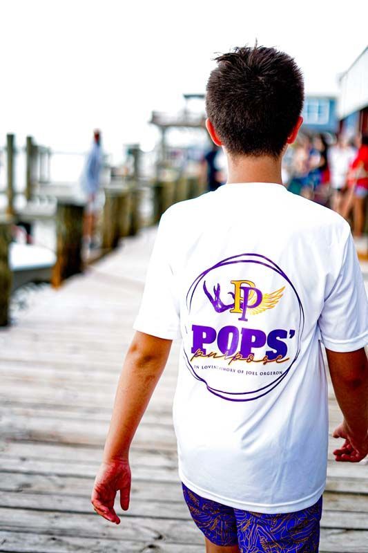 A young boy wearing a white t-shirt that says pops is walking on a pier.