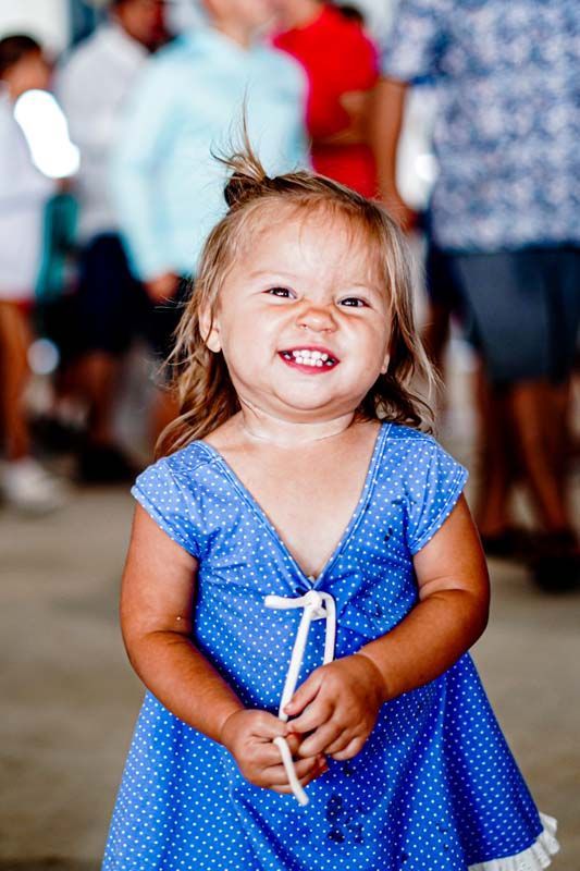 A little girl in a blue dress is holding a lollipop and smiling.