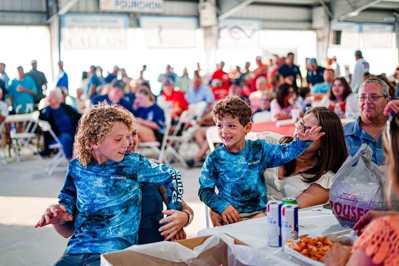 A group of children are sitting at a table in front of a crowd of people.