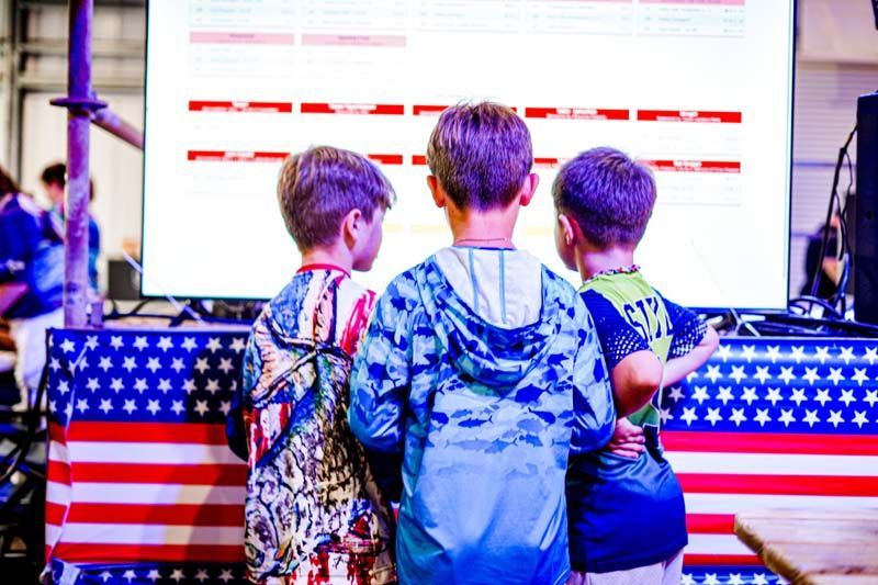 Three young boys are standing in front of an american flag and looking at a large screen.