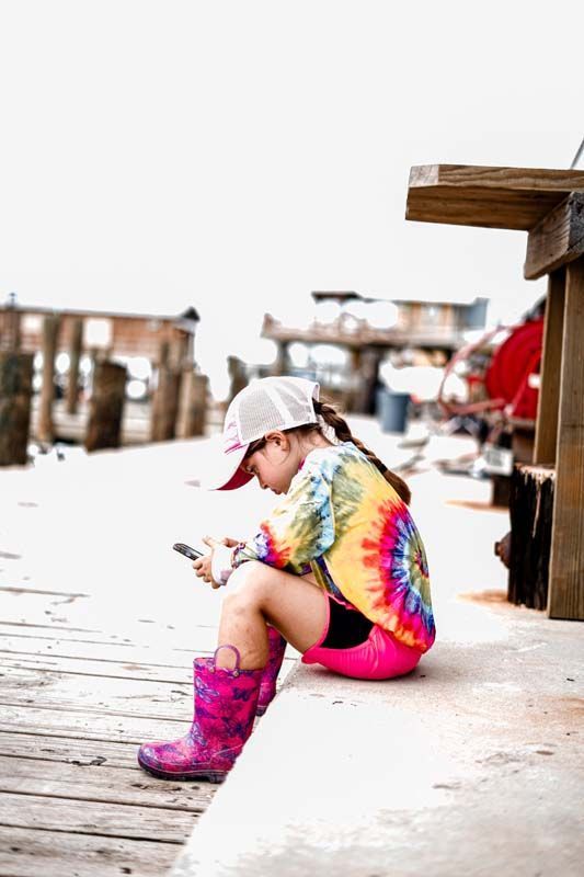 A little girl is sitting on a dock looking at her phone.