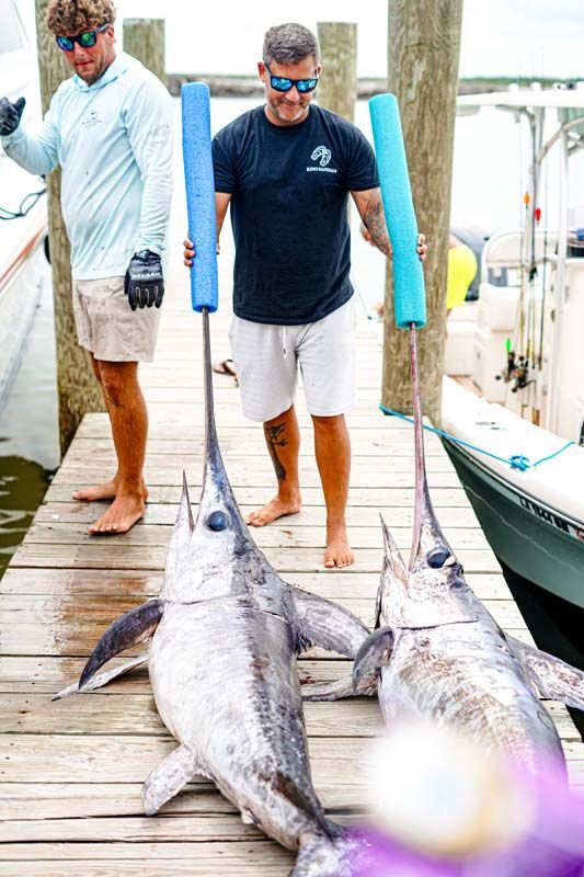 Two men are standing on a dock holding two large fish.