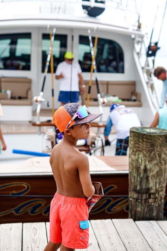 A young boy wearing a hat and sunglasses is standing on a dock next to a boat.